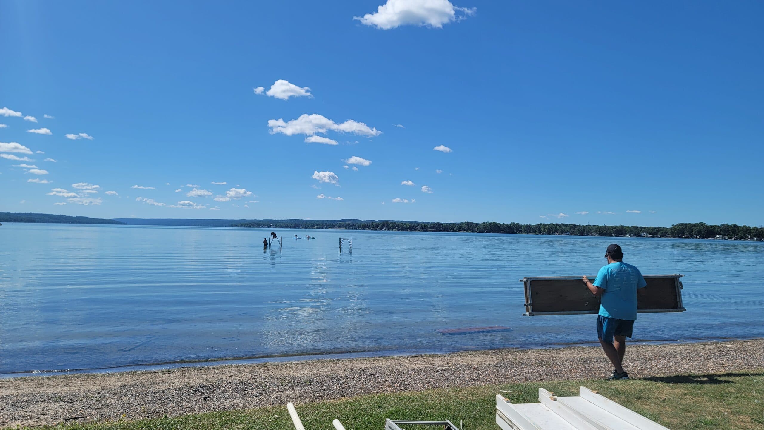 A Great Race volunteer carries a wooden dock section across the beach the day before the race, with Owasco Lake stretching out under a clear blue sky.