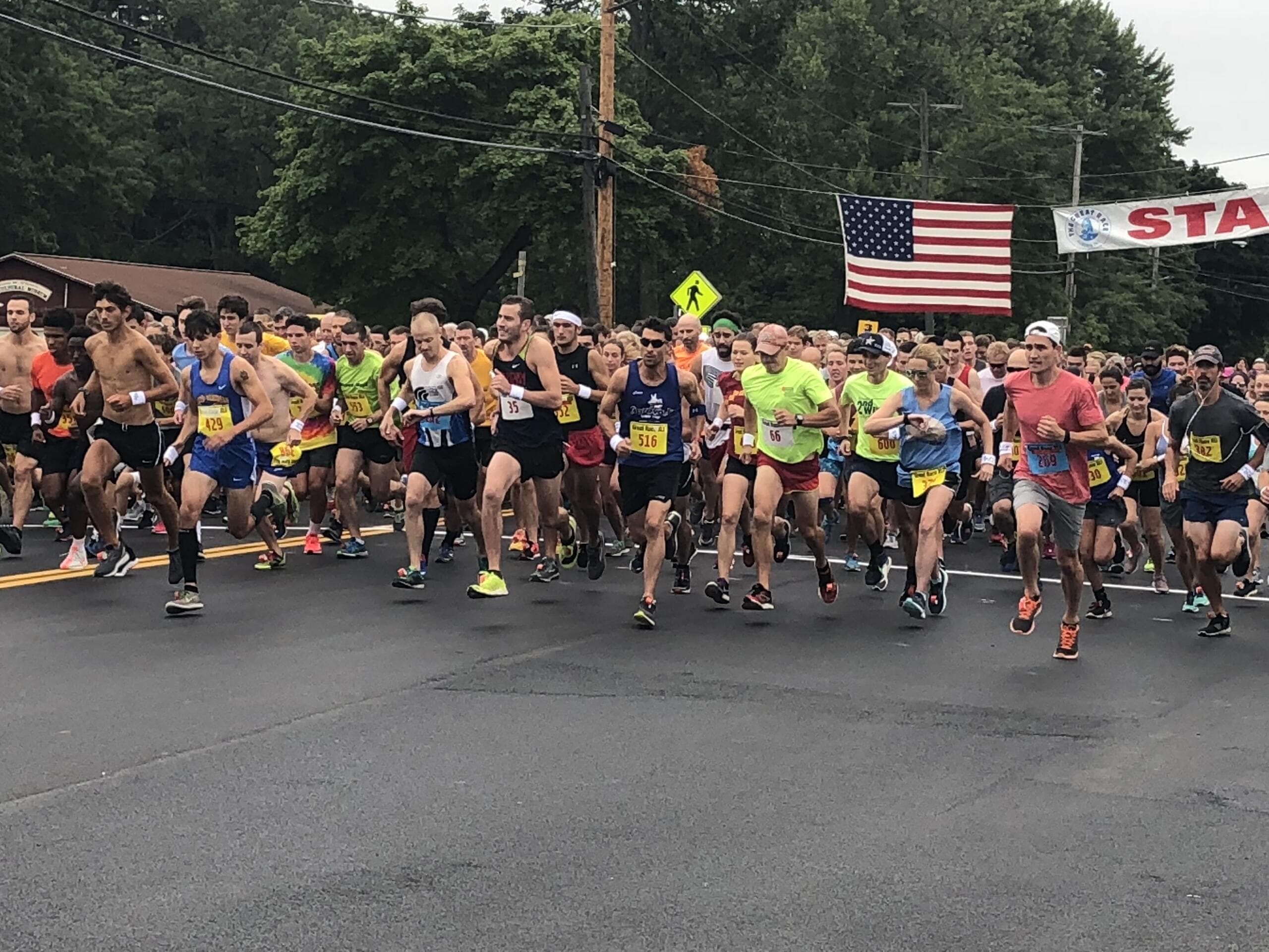 A pack of Great Race runners surging off the start line under the START banner and an American flag.